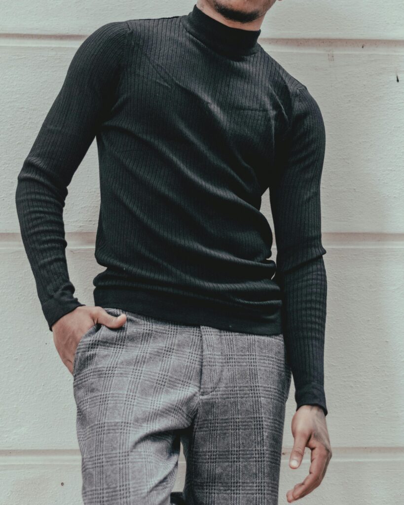 A fashionable man posing in a black turtleneck and plaid pants against a wall.