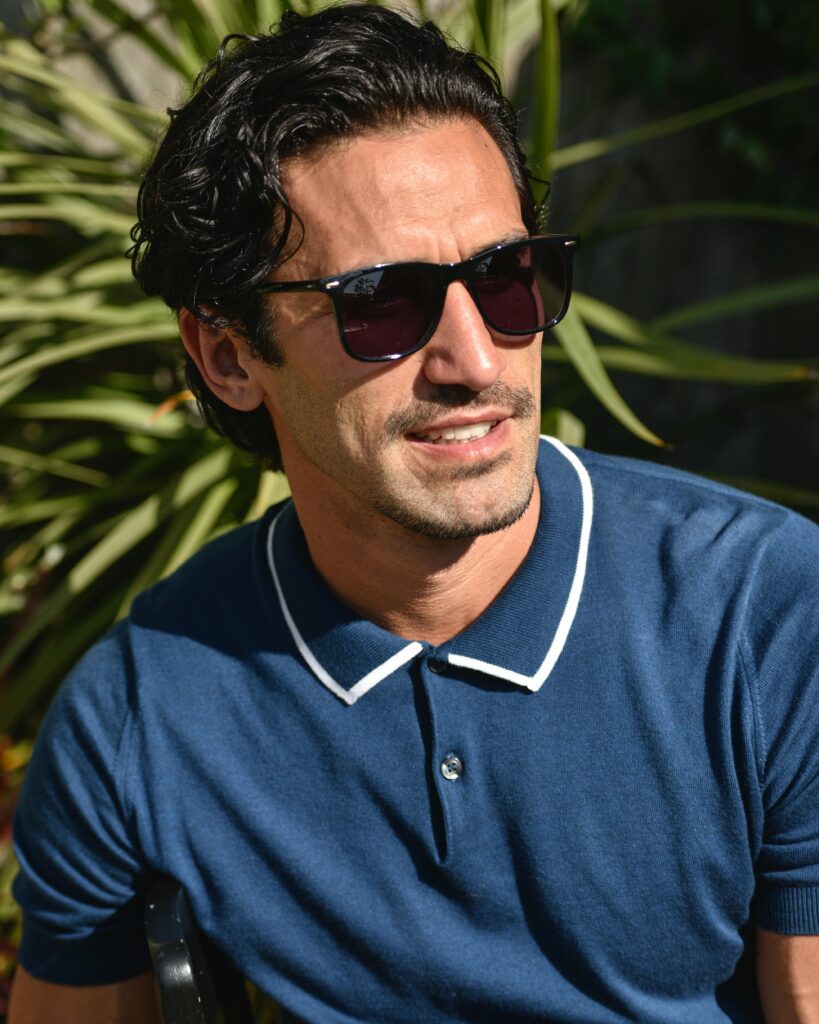 Confident man wearing sunglasses and polo shirt in bright sunlight outdoors.