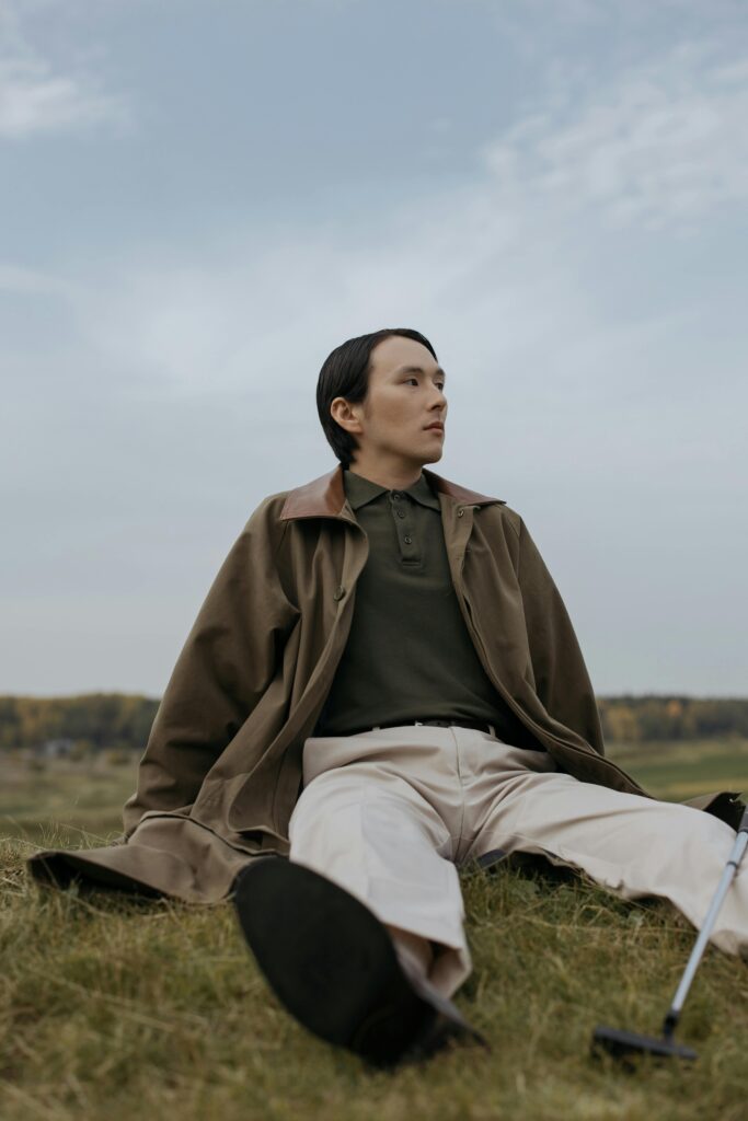 A man in a brown coat relaxing on grass in an outdoor setting, looking contemplative.