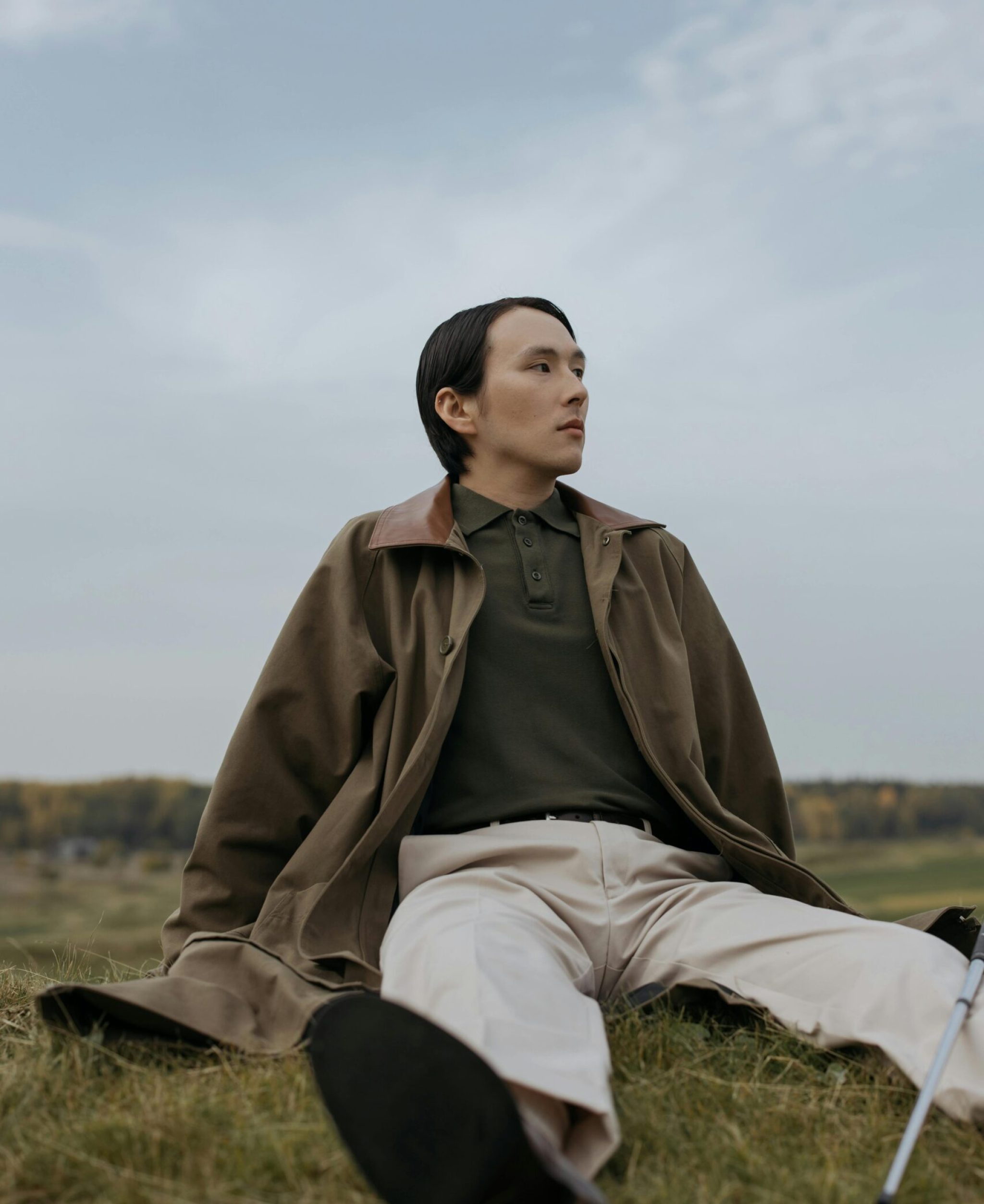 A man in a brown coat relaxing on grass in an outdoor setting, looking contemplative.
